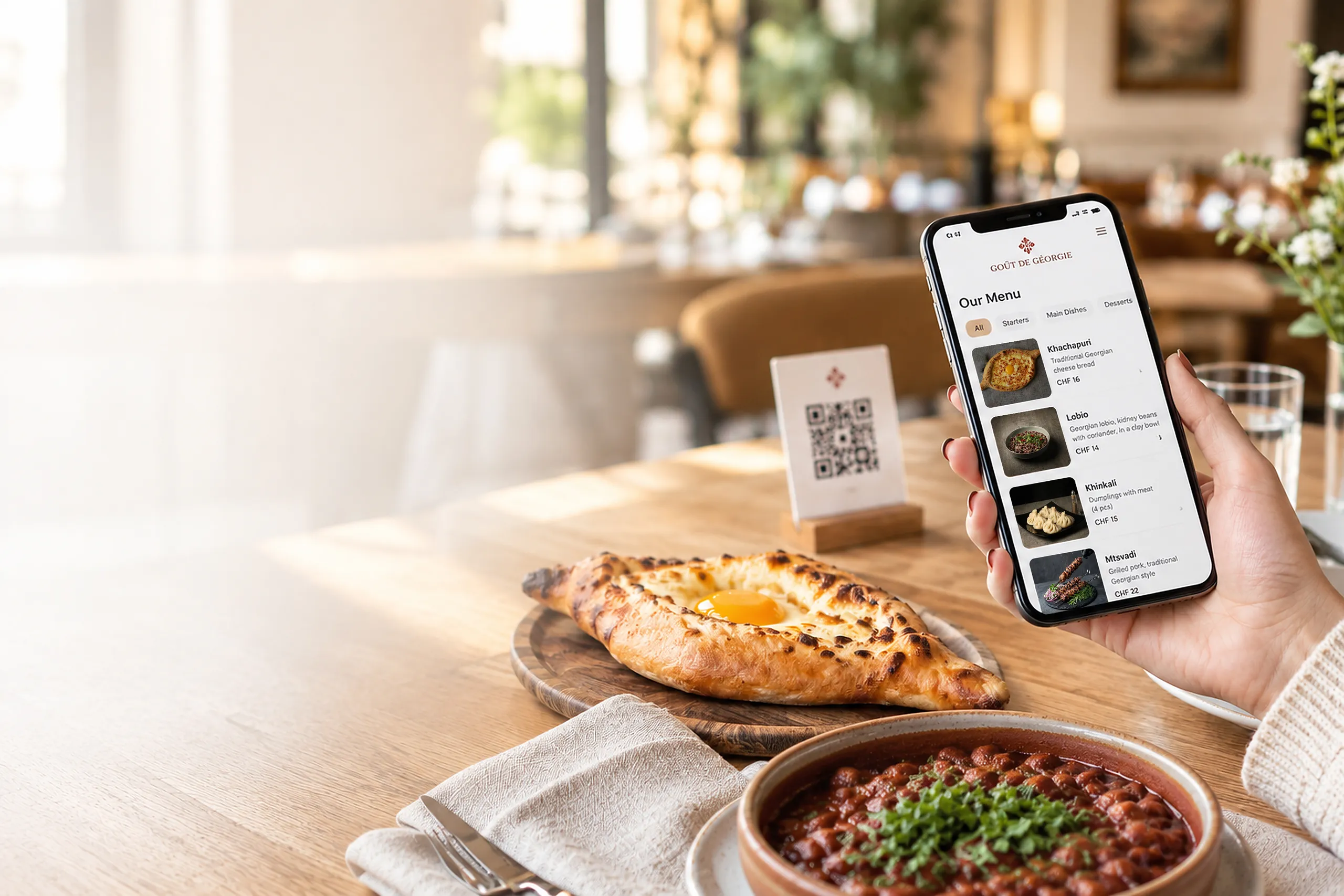 Cliente consultant le menu QR de Goût de Géorgie sur son téléphone, avec khatchapouri, lobio et présentoir QR sur la table